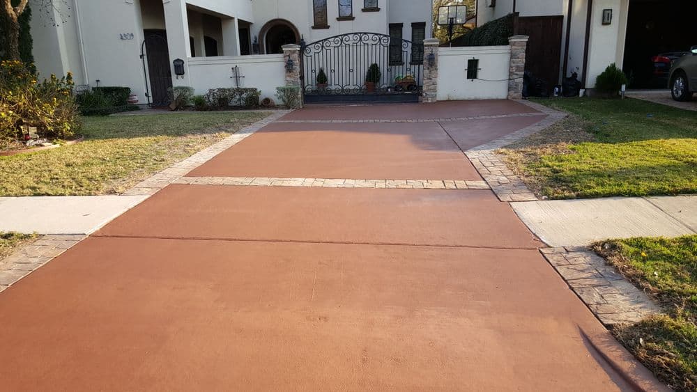 Newly poured decorative brown concrete driveway with stone border and lush green landscaping.